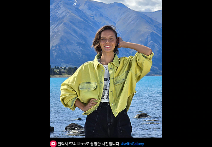 호숫가에서 포즈를 취하는 여성의 모습을 6.5배 줌으로 촬영한 사진입니다. 사진은 생동감이 넘치고 산 배경은 또렷하게 보입니다.